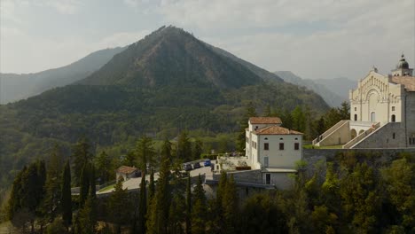 spectacular aerial dolly of mountaintop village and church, sanctuary of montecastello, with a mountainous and forest-covered background