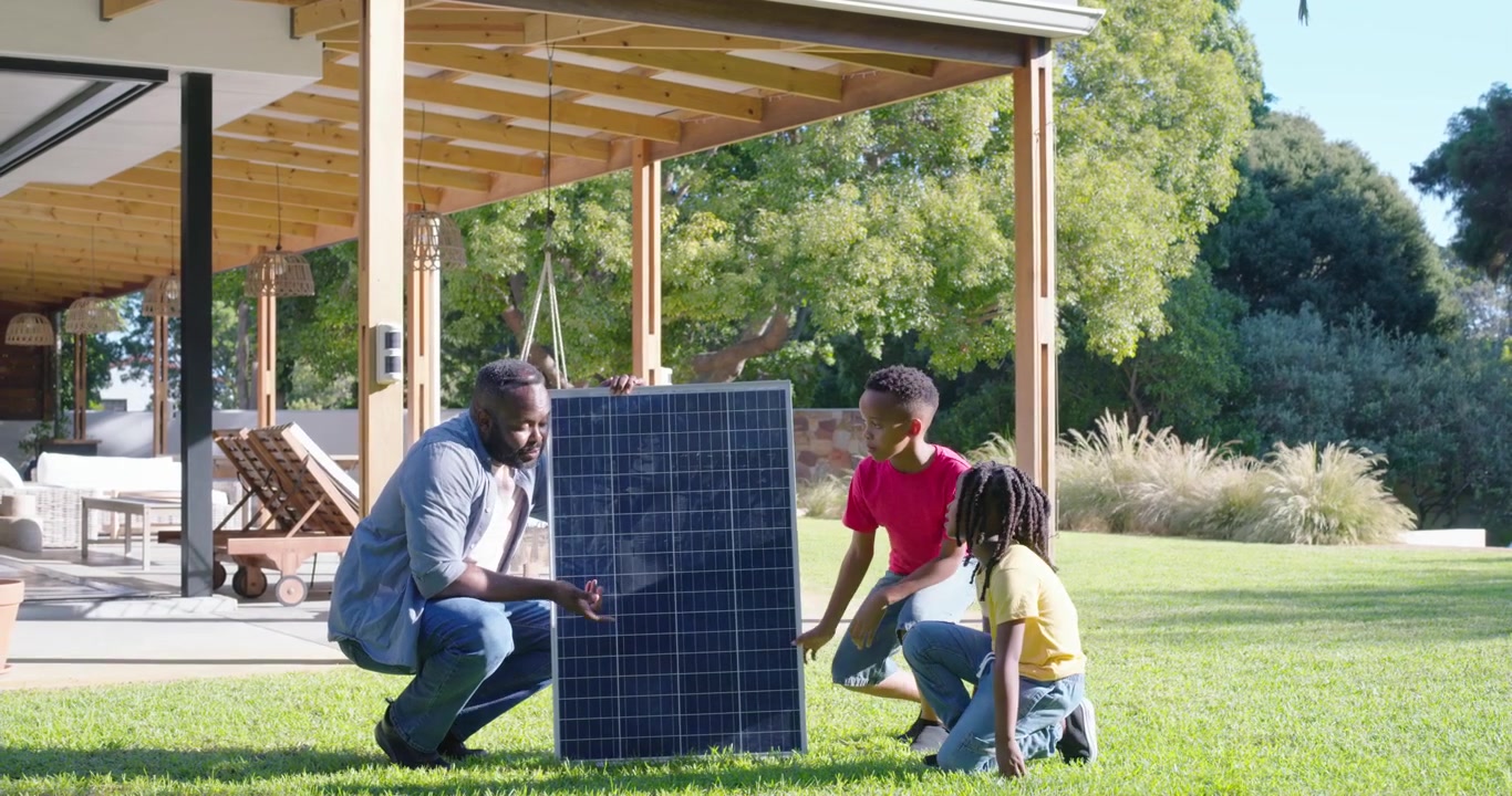 Afroamerikansk far i en familie bærer solpanel fra terrasse til græsplæne, mens solen skinner og han underviser børnene.