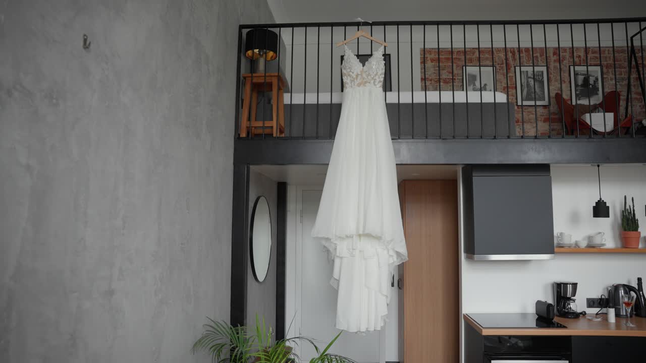 A beautiful wedding dress hangs gracefully from a railing in a stylish, modern loft apartment. Natural light highlights the intricate lace details, creating a serene ambiance