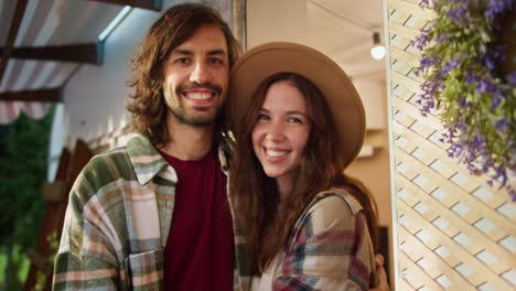 Close-up-portrait-of-a-happy-brunette-guy-with-stubble-and-a-plaid-shirt-and-a-red-T-shirt-who-stands-and-smiles-and-strokes-his-brunette-girlfriend-in-a-light-brown-hat-near-a-trailer-during-his-vacation-at-a-camp-during-a-picnic-outside-the-city-in-summer