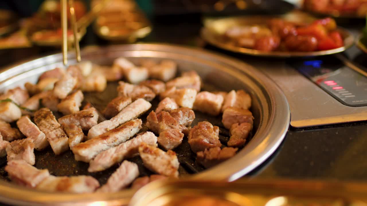 Sliced pork belly cooks on tabletop grill, golden lighting, close-up, chopsticks turning meat pieces