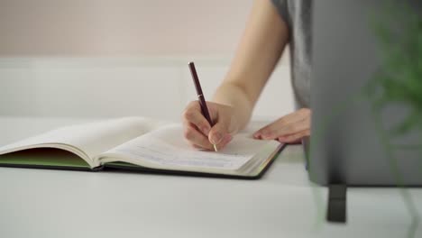 woman is writing down notes while working remotely as a digital nomad, using pencil and white paper book just near the laptop