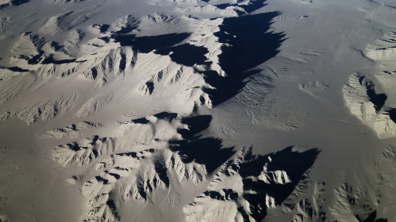 Drone che panoramica rivelando creste e guglie erose su un altopiano brullo, con un pennacchio di polvere sollevato da una raffica di vento.