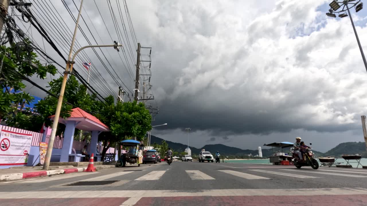 Low-angle view of cars and scooters on seaside street under dramatic storm clouds, natural daylight
