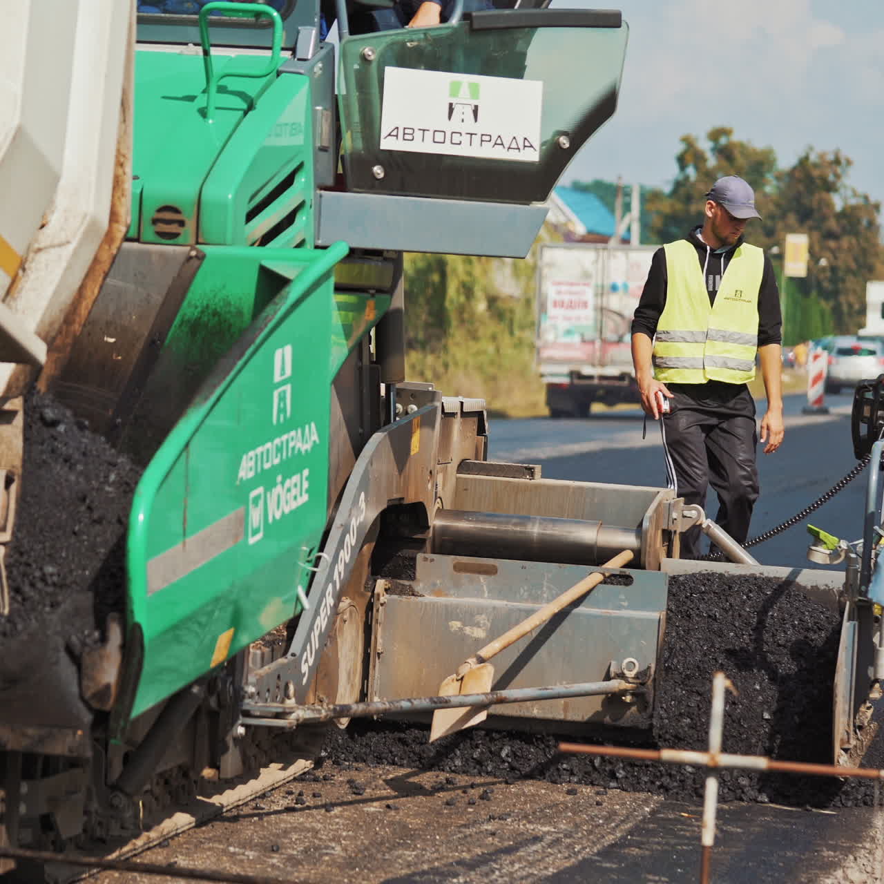 Big green paver machine pouring out asphalt and a worker on the road. Asphalt spreader at work in summer. Male worker with measuring tape during roadworks.