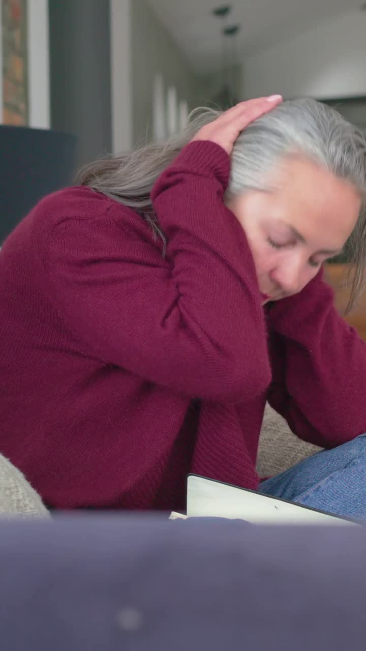 Video verticale: Donna che scrive con cardigan color bordeaux chiude il diario sul divano e si sfrega le tempie pensierosa.