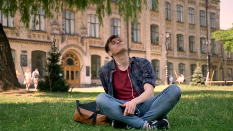 young handsome man is sitting on grass in park, listening music with earphones, relax concept, dreaming concept, building on background