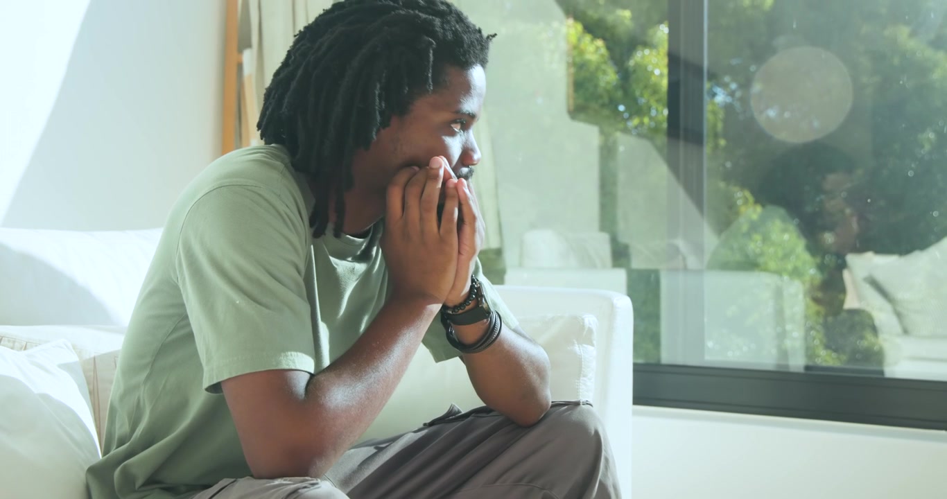 Homem afro-americano sentado no sofá perto da janela, vestindo uma camiseta, esfregando os olhos enquanto a luz do sol atrai seu olhar