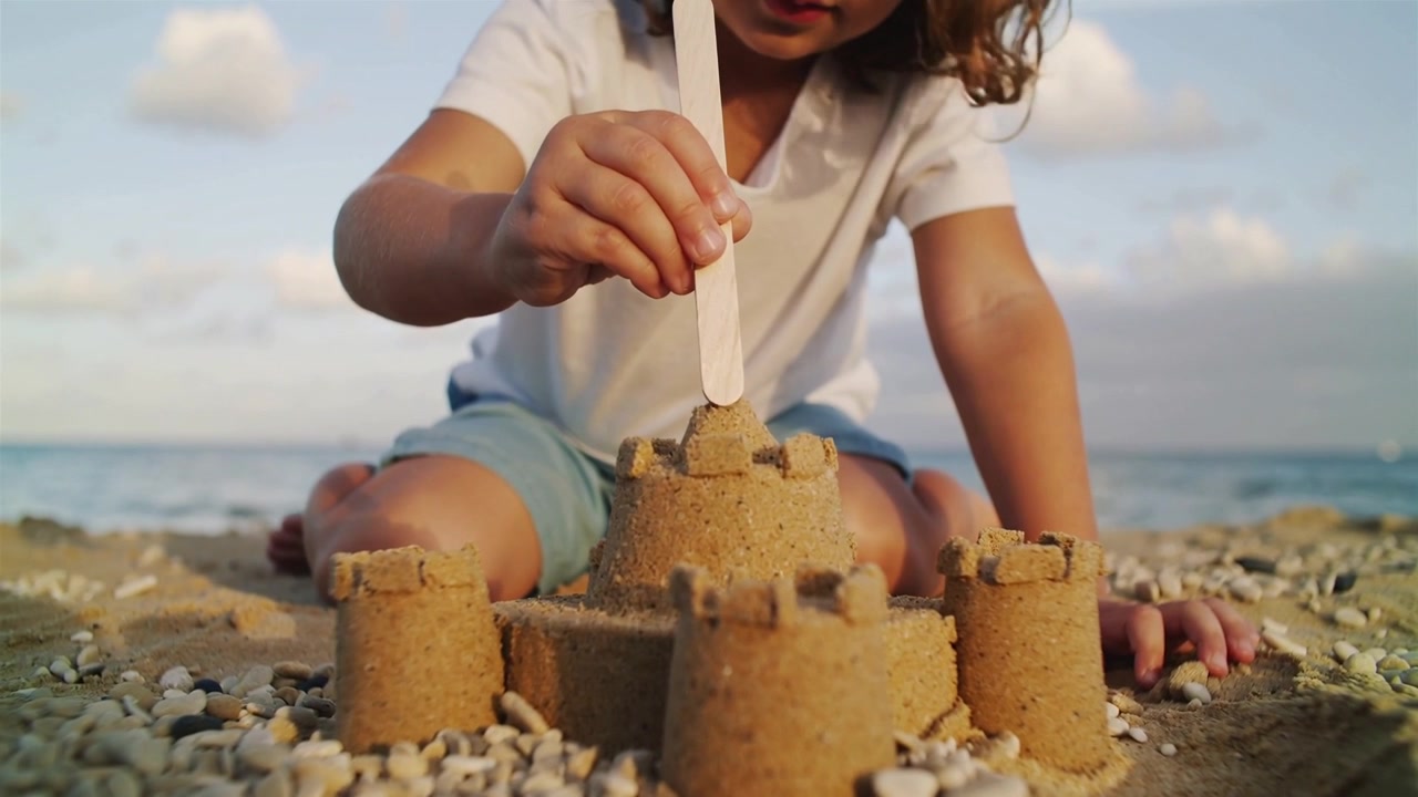 Menina de camiseta branca e shorts azul, com um graveto, moldando um castelo de areia na praia e finalizando a torre