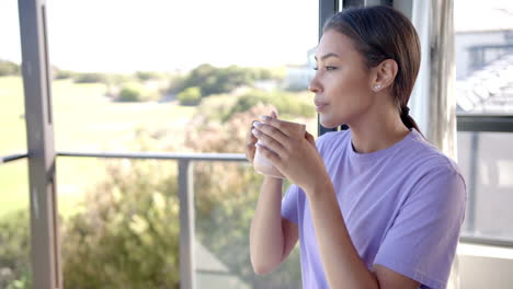 woman in purple shirt holding coffee cup, looking out window at home, copy space