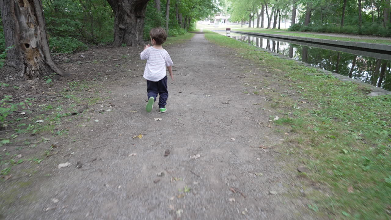 Child running in outdoor garden. Kid skipping and walking through the forest and walking beside duck pond. Camera moving forwards and following a happy young boy.