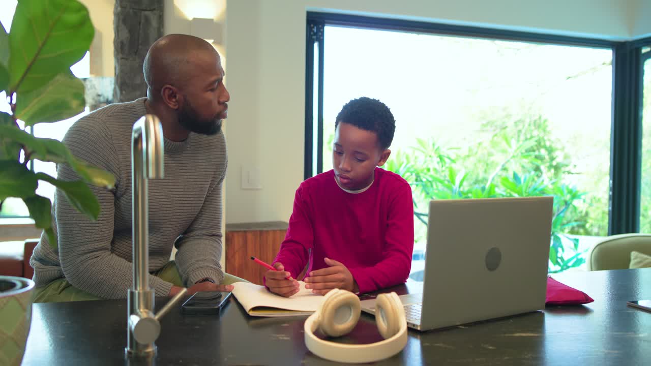 African American father and son opening notebook, studying at kitchen island, discussing homework