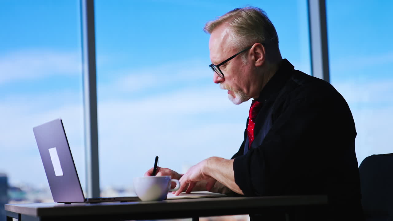 Busy Caucasian man wearing black shirt, vest and red tie works at desk. Man talks and writes down in his paper notebook.