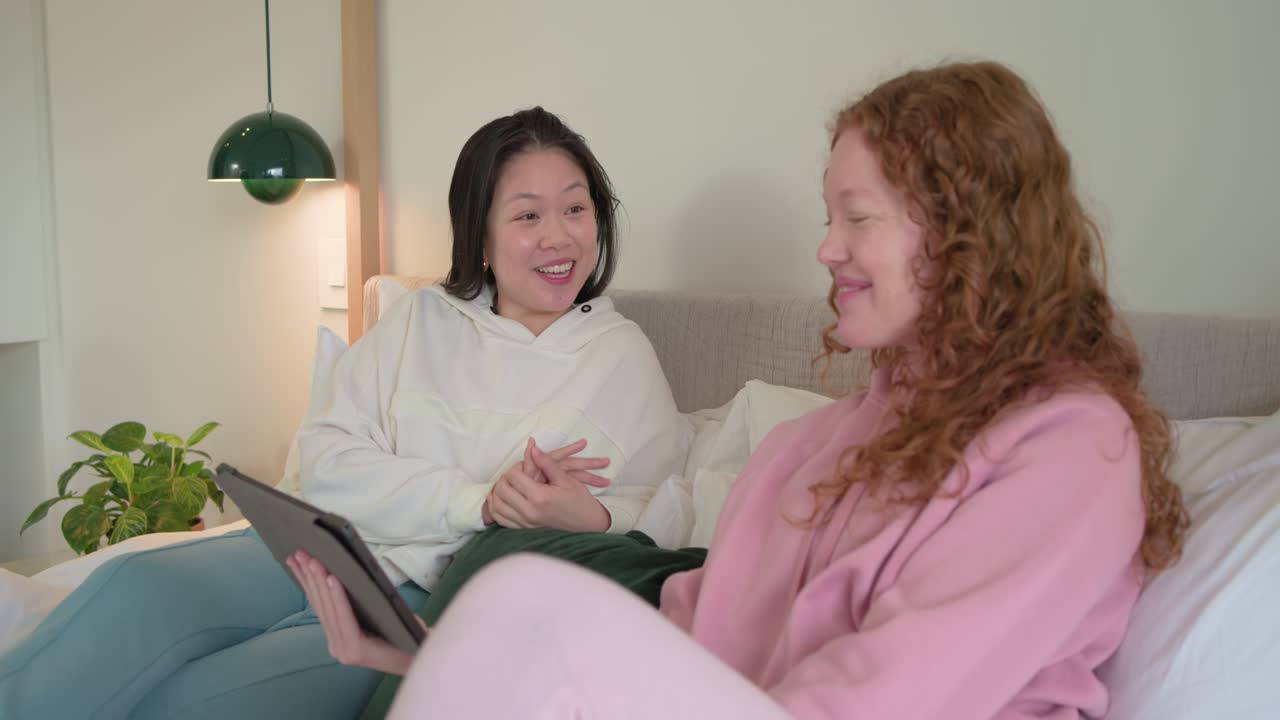 Due donne sedute a letto, una mostra una clip su un tablet, l'asiatica con la felpa bianca indica qualcosa, quella con la felpa rosa ride