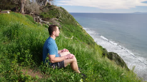 a fit strong man sitting in a meditating pose while doing deep breathing exercises on the edge of a beach cliff overlooking the ocean in santa barbara, california