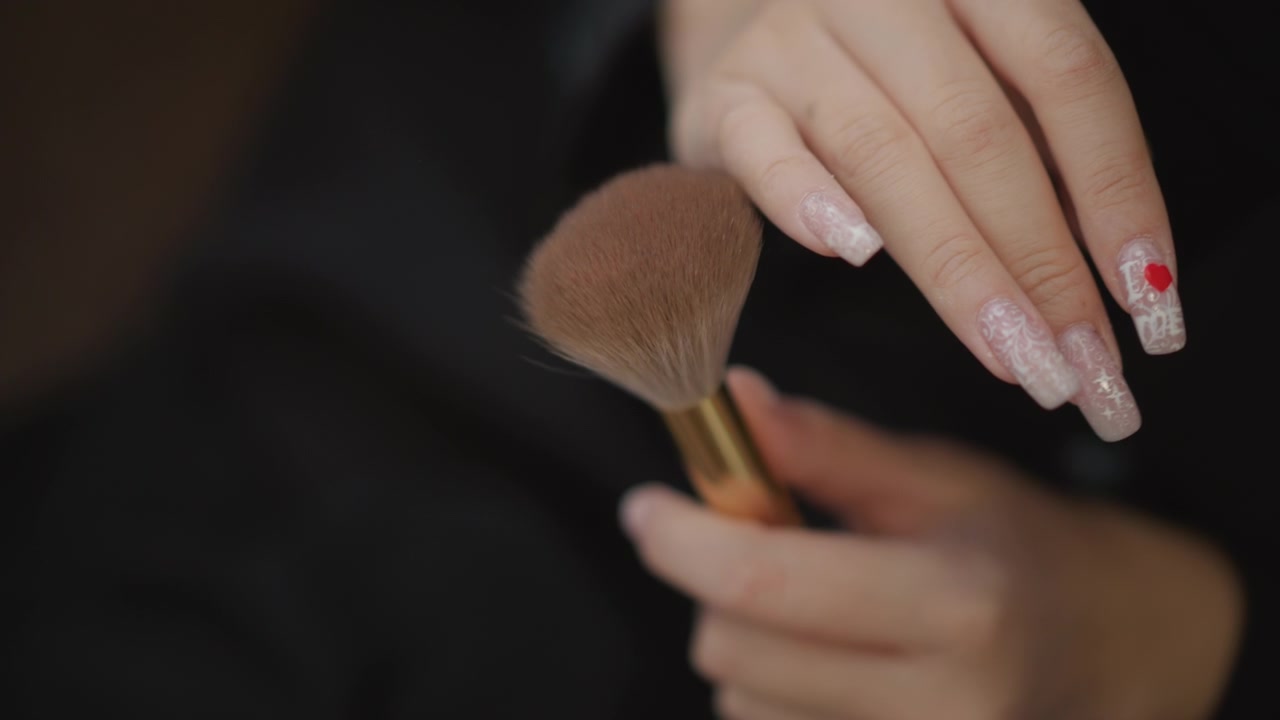 Close view of manicured hands holding makeup brush and gently tapping bristles against fingertips in soft light, creating calm focused beauty routine with delicate motion detail and refined texture