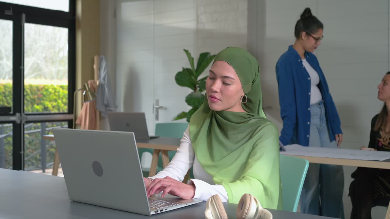 Seorang wanita berhijab mengetik di laptop di kantor, rekan kerja membawa kertas sambil mengobrol, ruang salin