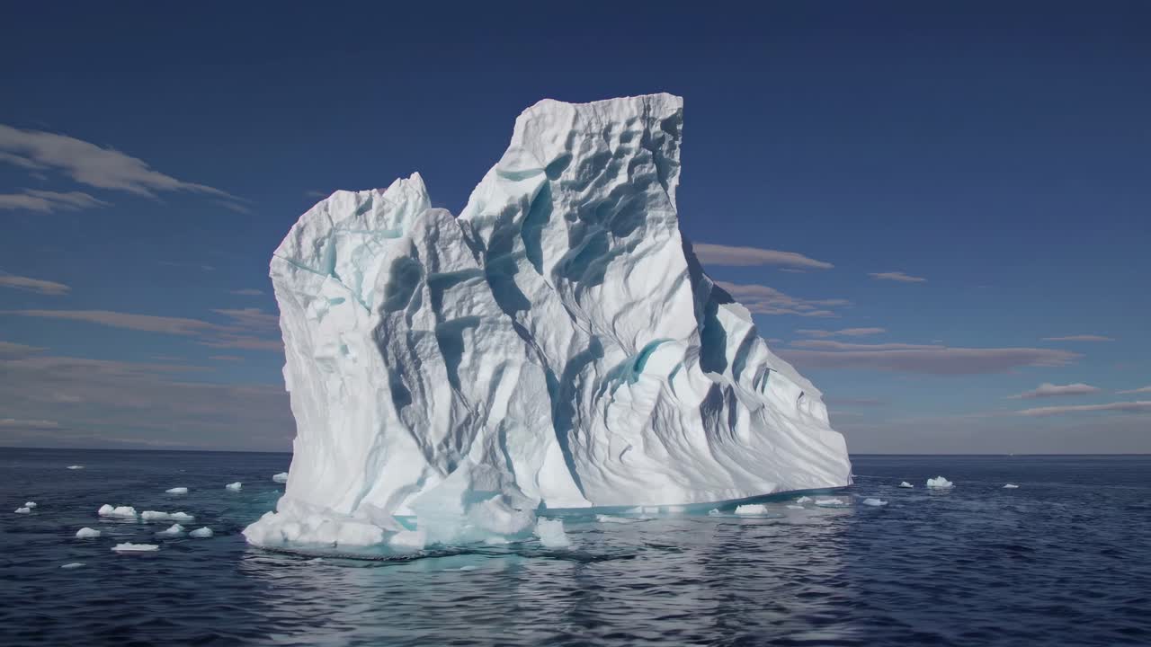 A majestic iceberg floats in calm waters under a clear sky. Captured from a low-angle, this video