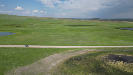 side tracking aerial view of car driving down countryside road passing lakes, green pastures, and cows