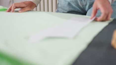 female tailor carefully places cut out paper design over light green elastic fabric on workspace table in sewing studio, preparing material layout for precise cutting and creative garment making