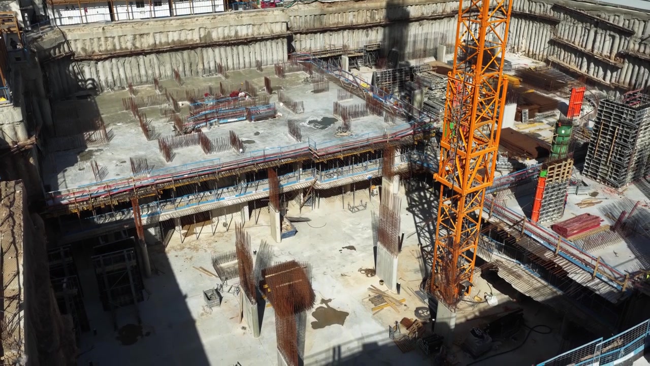 Aerial drone view of a large-scale construction site in Tel Aviv, showing an active tower crane and the structural foundation of a new skyscraper against the city skyline.