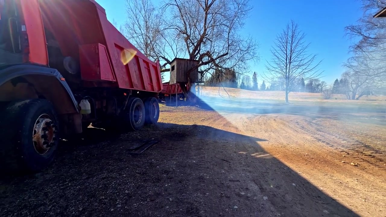 Red dump truck parked on gravel with exhaust smoke drifting in bright sunlight, capturing heavy-duty transport machinery in a rural industrial setting.