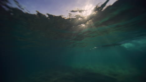 Shimmering-light-sparkles-across-underwater-ocean-surface-as-longboard-surfer-glides