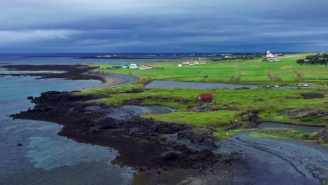 Vista-Aérea-De-La-Costa-Volcánica-Con-Campos-Verdes-Y-Casas-En-Gardakirkjugardur,-Gardabaer,-Islandia