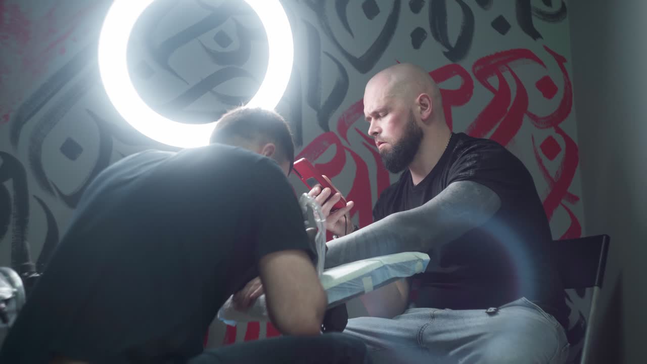 Tattooist making tattoo on arm of client with smartphone