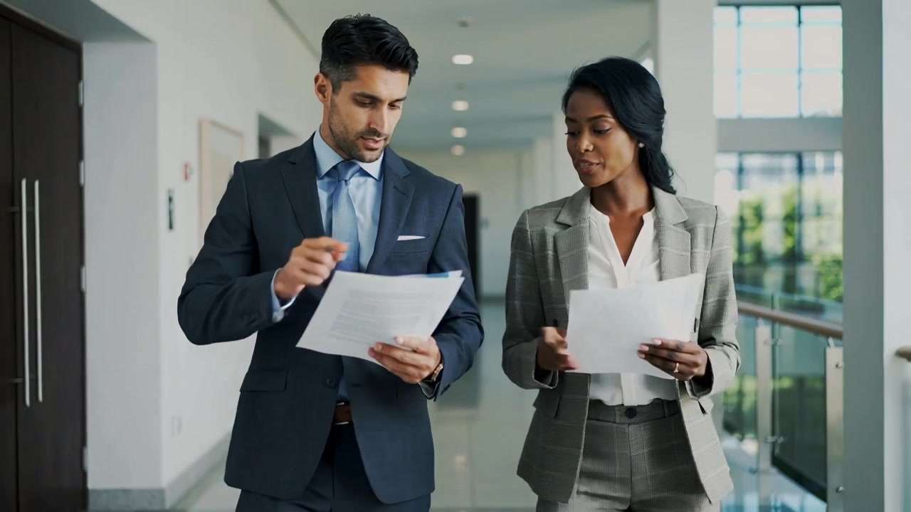 Walking and pointing, man in suit and woman in suit reviewing work documents in bright corridor