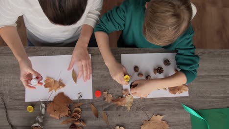 Mutter-Und-Sohn-Machen-Bilder-Von-Herbstblättern.Familien-,-Kreativitäts--Und-Bastelkonzept-–-Mutter-Und-Kleiner-Sohn-Machen-Mit-Klebestiften-Und-Papier-Zu-Hause-Bilder-Von-Trockenen-Herbstblättern,-Tannenzapfen-Und-Kastanien