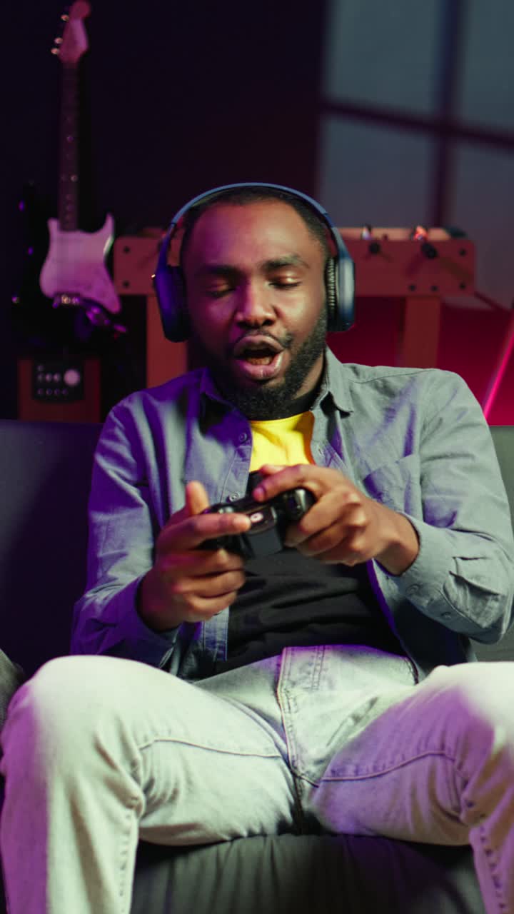 Vertical video Enthusiastic gamer on couch using controller and headphones to play videogame