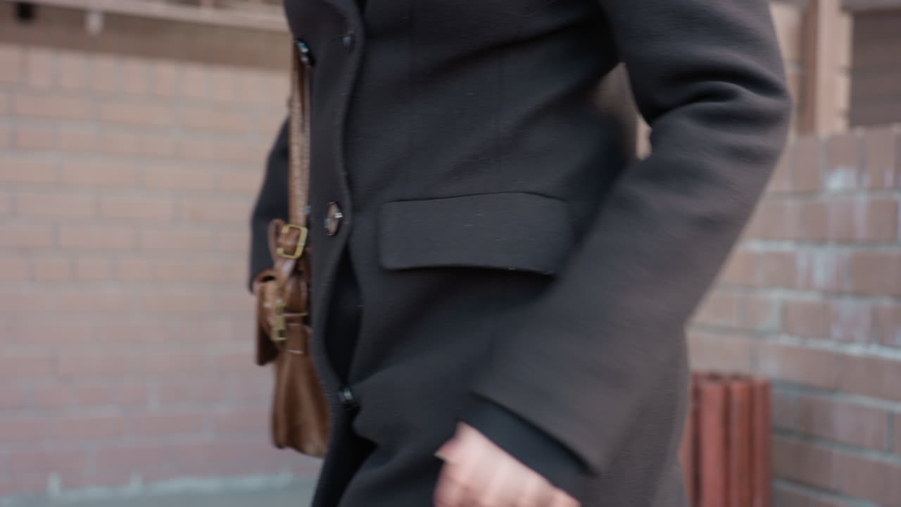 옆모습은 검은색 모직 정장을 입은 젊은 여성이 벽돌 벽을 지나가는 모습을 보여줍니다. 손은 갈색 가죽 가방을 안정적으로 잡고 있으며, 미묘한 움직임은 깨끗한 직물 질감으로 시원한 날씨에 걷는 모습을 암시합니다.