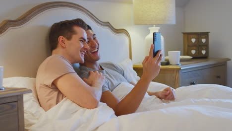 Pareja-Masculina-Del-Mismo-Sexo-Acostada-En-La-Cama-En-Casa-Haciendo-Videollamadas-En-El-Teléfono-Móvil