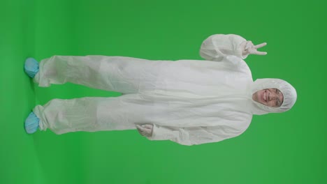 full body of asian male wearing personal protective equipment uniform ppe smiling to camera with a victory hand sign in green screen studio