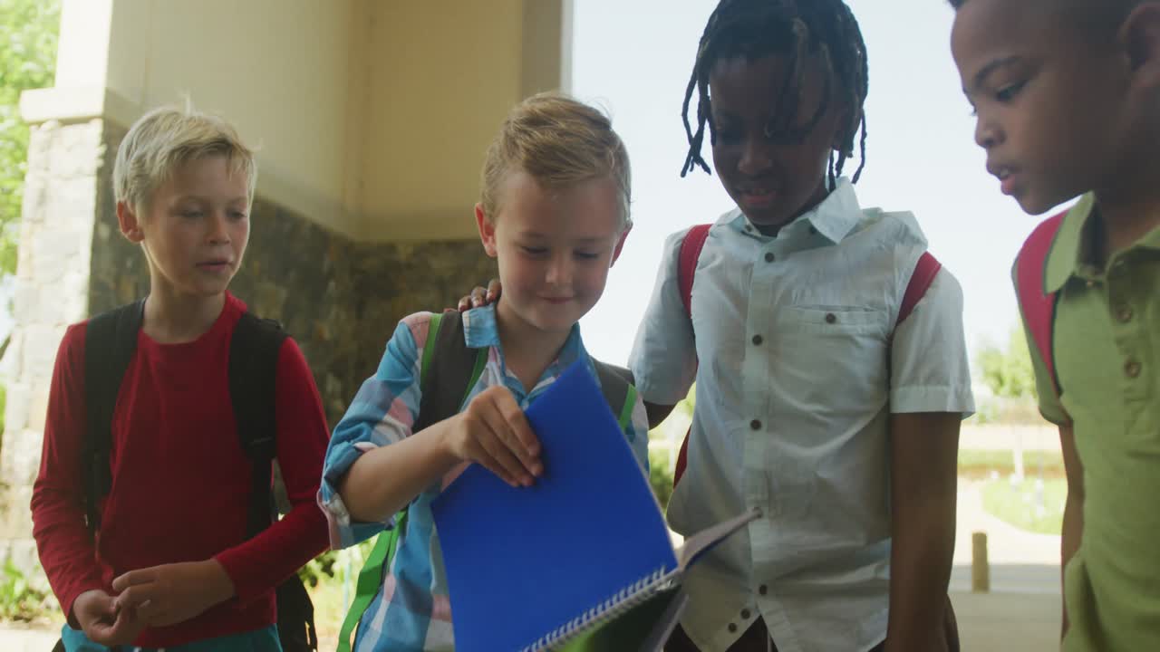 Video of happy diverse boys walking, looking at notebook in front of school