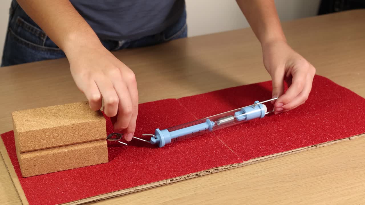 A hand pulls a wooden block across sandpaper using a spring scale, demonstrating friction and force measurement