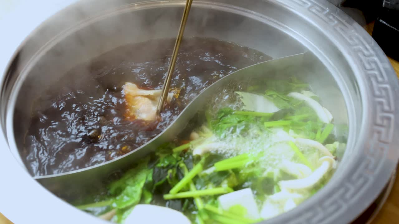 Chopsticks cook sliced meat in steaming dual broth hotpot with fresh vegetables, overhead view