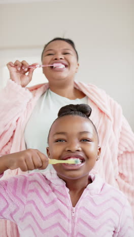 vertical video of happy african american mother and daughter brushing teeth, in slow motion