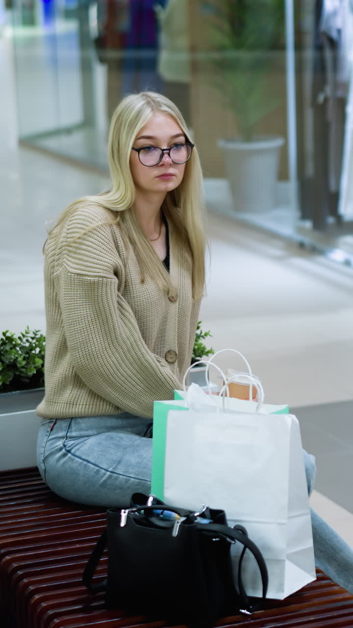 장식용 식물 근처에 앉아있는 전문적인 여성이 고민스럽게 커피를 마시며, 배경에는 마네킨과 지나가는 쇼핑객이 있는 옷 가게가 있으며, 현대적인 쇼핑 분위기를 만니다.