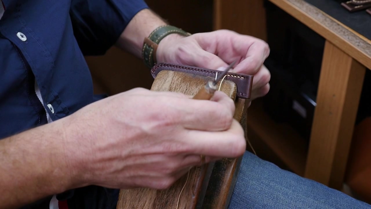 Primo piano delle mani di un artigiano che cuciono un cinturino personalizzato in pelle marrone con filo cerato e punteruolo su un tradizionale morsetto da cucito in legno in un laboratorio.