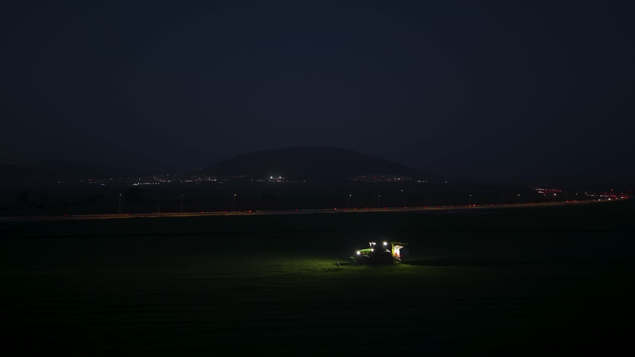 Il lavoro agricolo notturno si svolge in una vasta valle all'ombra di lontane cime scure con le luci dei trattori che trafiggono la notte.