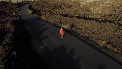 a woman with a fluttering dress in the wind stands in the middle of a road built on a volcanic landscape, hierro island, canary - aerial circle shot