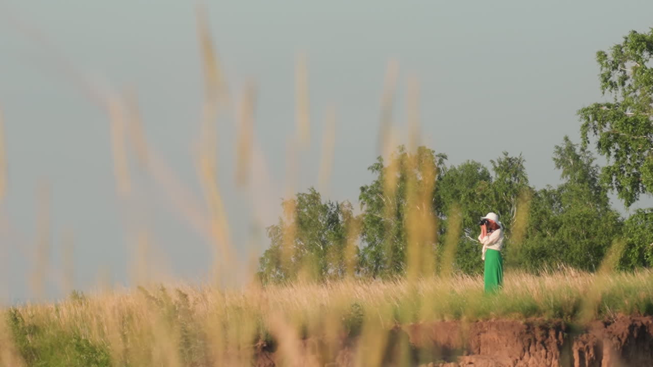 distant view of solo adventurer in white hat and green skirt photographing landscape near cliff edge surrounded by tall grass and vibrant trees under clear blue sky