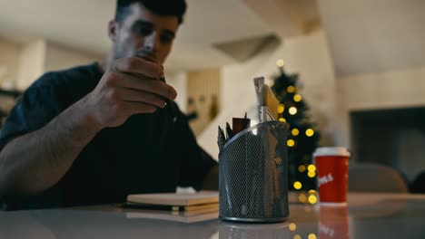 una foto de perfil capturando al artista mientras selecciona lápices de un soporte de malla de alambre, en el telón de fondo de un árbol de navidad fuera de enfoque y luces de hadas bokeh