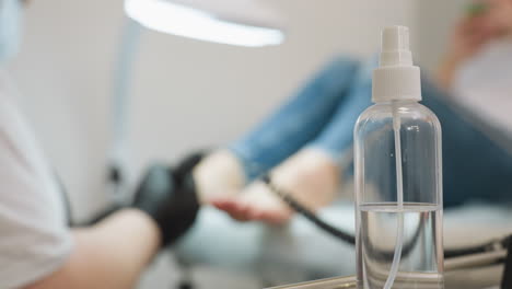 Clear spray bottle positioned in foreground on salon table while blurred view in background shows nail technician wearing gloves and mask filing client toenails under bright lamp during pedicure