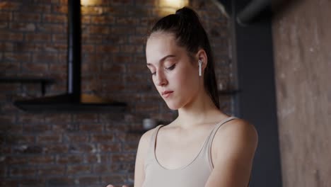 a young woman in a tank top and headphones stands in a kitchen and checks her watch.