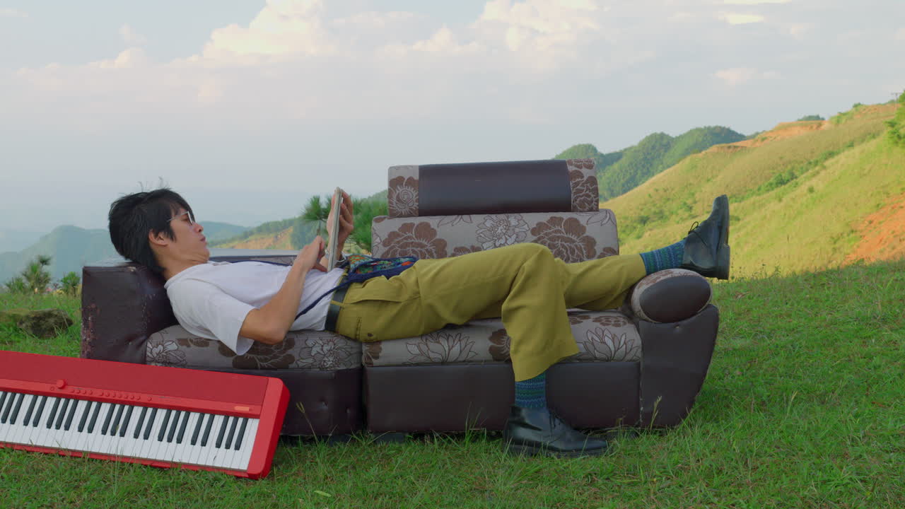 young asiatic artist laid on a couch outdoor in valley mountains , musician reading a book with a a piano keyboard finding inspiration for a new song