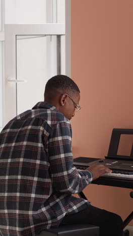 man in glasses enjoys playing synthesizer. black performer rehearses melodic sequence on electric instrument alongside bookshelf. rehearsal at home