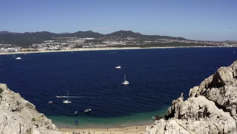 Yachts-moored-in-ocean-shallows-with-Cabo-San-Lucas-beach-aerial-reveal,-4K
